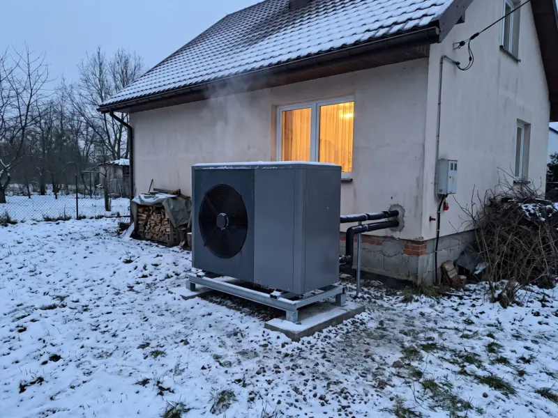 Pompa ciepła powietrze-woda w ogrodzie zimą, z jednostki unosi się para, w oknie domu ciepłe światło