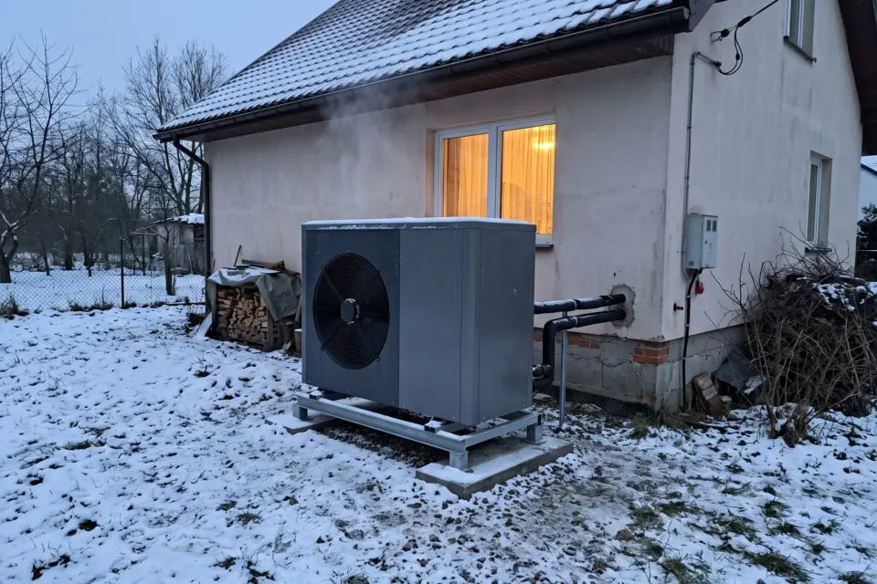 Pompa ciepła powietrze-woda w ogrodzie zimą, z jednostki unosi się para, w oknie domu ciepłe światło